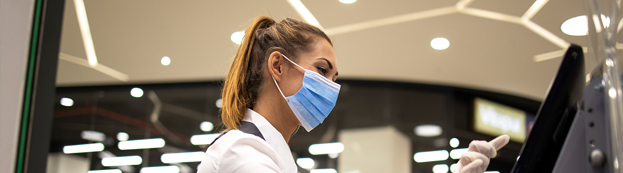 A female Needs employee is serving customers at the cashier while wearing her hygienic face mask.